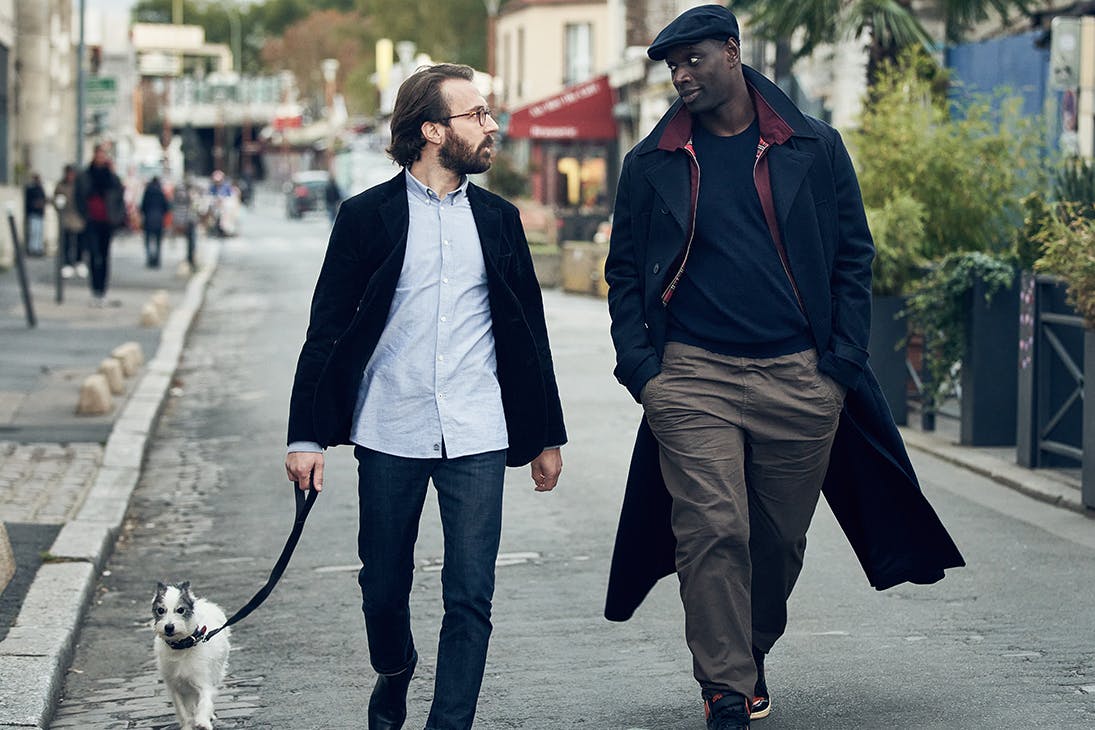 Two men, one holding a dog's leash, walk down a village street.